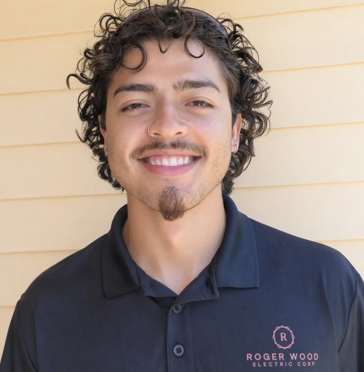 Man with curly dark hair and facial hair smiling at camera wearing navy Roger Wood Electric Corp polo shirt